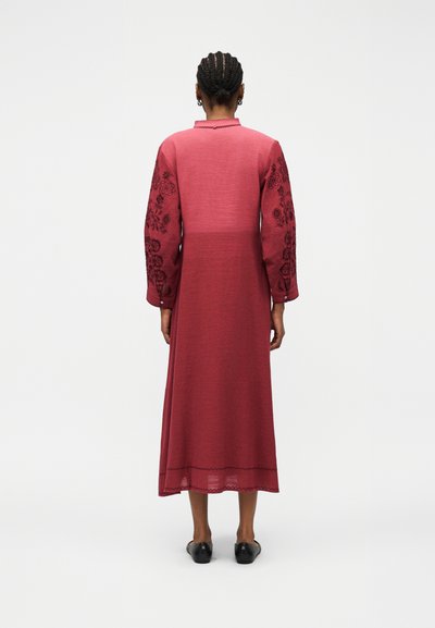 Γυναίκα με πλεξούδες στα μαλλιά που φοράει μακρύ κόκκινο φόρεμα με κεντήματα λουλουδιών στα μανίκια, στέκεται με την πλάτη προς τον θεατή.