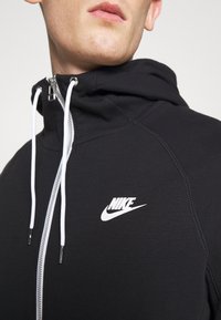 Svart zip-up hoodie tillverkad av mjukt tyg, med vita dragsnören och en framträdande vit Nike-logotyp på bröstet.