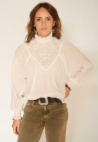 Blouse blanche légère avec des manches ballons longues, un col haut à volants et une broderie florale sur le buste. Assortie avec un jean marron et une ceinture noire.