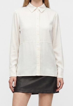 Femme portant un chemisier blanc à manches longues boutonné et une mini-jupe en cuir noire, debout devant un fond uni.