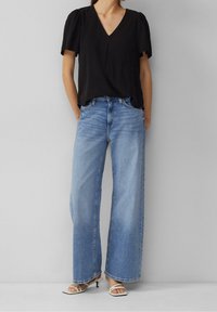 Top nera a maniche corte con scollo a V abbinato a jeans a vita alta di colore blu chiaro e scarpe con cinturini bianchi su uno sfondo neutro.