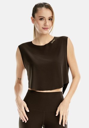 Junge Frau mit zurückgestrichenen Haaren, die ein ärmelloses schwarzes Croptop und hoch taillierte schwarze Hosen trägt, steht vor einem weißen Hintergrund.