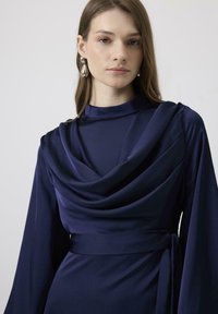 Rochie din satin de culoare albastru închis, cu decolteu drapat, mâneci lungi și un detaliu la talie legat. Textură netedă și siluetă elegantă.