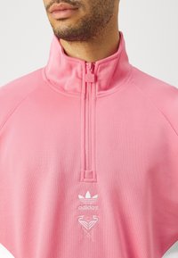 Gaiši rozā Adidas džemperis ar augstu apkakli, ceturtdaļas rāvējslēdzēju un baltu logotipu uz krūškurvja, izgatavots no mīksta, teksturēta auduma.