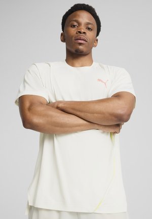 T-shirt de sport crème clair à manches courtes, avec un logo Puma rose et de subtils accents jaunes le long des coutures. Tissu doux et lisse.