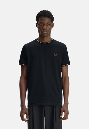 Hombre con una camiseta negra de manga corta con un pequeño logo de corona de laurel naranja en el pecho, de pie frente a un fondo claro y liso.
