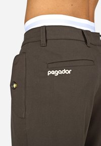 Zbliżenie na osobę w ciemno-brązowych spodniach, z widocznym białym pasem i haftem "pegador" w pobliżu tylnej kieszeni.