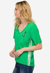 Grøn bomulds T-shirt med V-hals, med et anker grafik på fronten og et lysere mønstret sidepanel. Kortærmede, afslappet pasform.