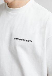 Weißes Baumwoll-T-Shirt mit runder Ausschnitt, kurzen Ärmeln und "PROHIBIERT" in fetten schwarzen Buchstaben auf der linken Brustseite.