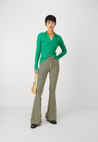 Top verde texturat, cu mânecă lungă și decolteu în V, purtat cu pantaloni evazați cu dungi negre și galbene, cu talie înaltă. Accesoriile includ o geantă mică.