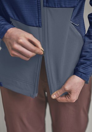 Hände, die einen grauen Reißverschluss an einer blauen Jacke hochziehen, die über braunen Hosen getragen wird, mit Fokus auf den Reißverschluss und die Stoffdetails.