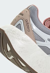 Le sneakers Adidas Adizero presentano una tomaia in mesh grigia con dettagli rosa, una intersuola bianca e un design della suola in gomma testurizzata.