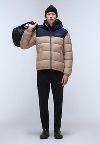 Giubbotto puffer beige e blu navy con cuciture orizzontali, chiusura con zip e cappuccio. Berretto nero e borsa; pantaloni e scarpe nere.