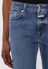 Gros plan d'une personne portant un jean en denim bleu avec poches visibles, passants de ceinture, bouton, et main aux ongles peints reposant à côté de la jambe.