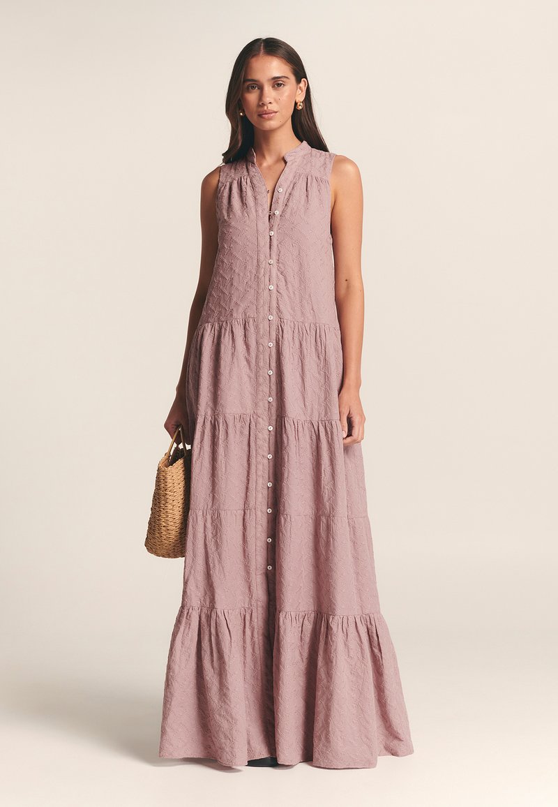 Robe longue sans manches avec des boutons de couleur mauve clair. Présente un design en couches, un tissu texturé et un col. Accessoirisée avec un sac tissé.