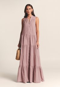 Robe maxi en lin de couleur mauve doux, avec un devant boutonné, un design sans manches, un ourlet à volants et un motif texturé subtil. Sac tissé en accessoire.