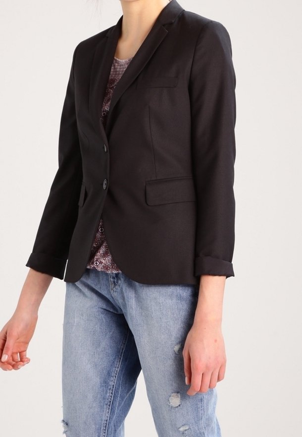 Personne portant un blazer noir ajusté sur un haut à motifs, associé à un jean bleu clair usé, debout devant un fond uni.