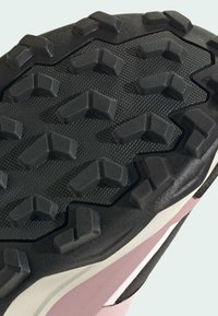 Μαύρη λαστιχένια εξωτερική σόλα με βαθιές, γωνιακές, σχήματος V ενδείξεις πέλματος σε ροζ και λευκή αθλητική σόλα, σχεδιασμένη για πρόσφυση.