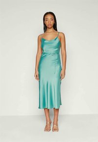 Ένα τιρκουάζ σατέν φόρεμα slip με λαιμόκοψη cowl, λεπτούς ρυθμιζόμενους ιμάντες και λεπτομέρεια με κόμπο στη μέση, που φτάνει κάτω από το γόνατο.