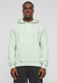Felpa verde menta realizzata in un materiale morbido, con tasca frontale, lacci e un piccolo logo sul petto. Jeans neri visibili sotto.