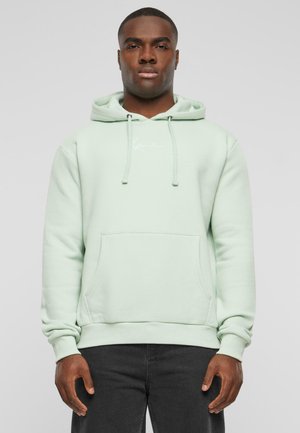 Mintgroene hoodie van een zacht materiaal, met een voorzak, koordjes en een klein logo op de borst. Zwarte jeans zichtbaar eronder.