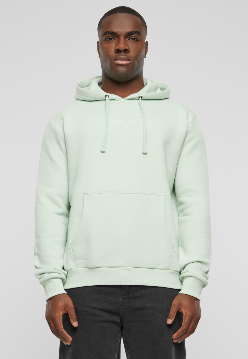 Felpa verde menta realizzata in un materiale morbido, con tasca frontale, lacci e un piccolo logo sul petto. Jeans neri visibili sotto.