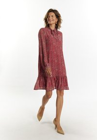 Rochie roșie cu imprimeu floral, cu hem ruffled, mâneci lungi și gât înalt cu un detaliu legat. Țesătura pare ușoară și texturată.