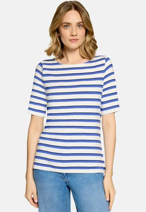 Femme aux cheveux blonds mi-longs portant une chemise blanche à rayures horizontales bleues et un jean bleu clair sur un fond uni.