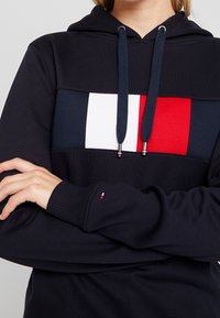 Námořnická mikina s kapucí s horizontálním blokovým designem, který obsahuje červené, bílé a modré sekce. Obsahuje malé logo na rukávu.