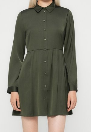 Dunkelgrünes, langärmeliges, knöpfbares Kleid mit Kragen, tailliertem Schnitt und ausgestelltem Rock, getragen von einer Person mit heller Haut und hellblonden Haaren.