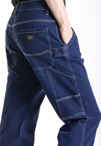 Pantalons cargo en denim bleu foncé, dotés de coutures doubles, de poches avant et arrière, et d'un patch logo jaune à l'arrière.