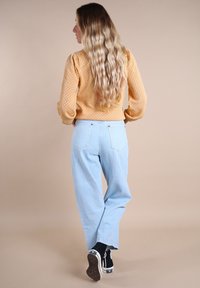 Un jean baggy bleu clair associé à un pull texturé de couleur moutarde. La tenue présente une coupe décontractée et des cheveux longs et ondulés.