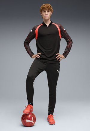 Jeune personne en tenue de sport noire et rouge, debout avec assurance, un pied posé sur un ballon de football rouge Puma, sur un fond gris.