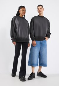 To personer iført uformell bekledning: en mørkegrå oversized hettegenser, svarte vide bukser, og en denim shorts, kombinert med svarte joggesko.