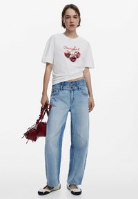 White graphic t-shirt featuring red cherries and "Desigual." Light blue loose-fit jeans with a faded finish. Red handbag and black-and-white shoes.