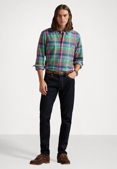 Homme aux cheveux mi-longs portant une chemise à carreaux verte et violette, un jean foncé, une ceinture marron, une montre-bracelet et des chaussures à lacets marron, se tenant avec une main dans la poche.