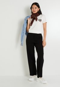 T-shirt blanc avec motif de cœur léopard, pantalon noir à jambes larges, veste en jean et écharpe à motifs bruns. Porté avec des baskets compensées blanches.