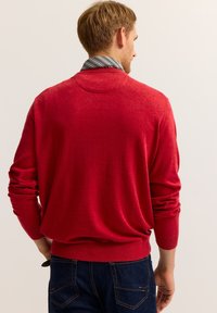 Roter Strickpullover mit rundem Ausschnitt und langen Ärmeln. Weiche Textur, gerippte Bündchen und Saum. Wird über einem gestreiften Hemd mit Kragen getragen.