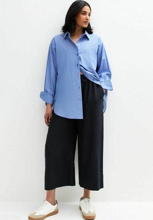 Camicia azzurra chiara button-up con taschino, vestibilità comoda e maniche arrotolate, abbinata a pantaloni neri larghi e sneakers bianche.