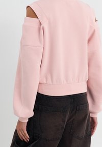 Pink sweatshirt with cut-out shoulders, textured fabric, and ribbed hem. Paired with dark, loose-fitting cargo pants featuring back pockets.