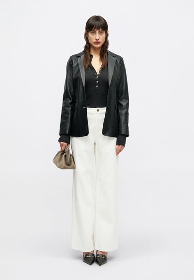 Donna che indossa un blazer di pelle nera, un top nero con bottoni, pantaloni bianchi a gamba larga, scarpe nere a punta, mentre tiene una borsa clutch beige, in piedi contro uno sfondo bianco.