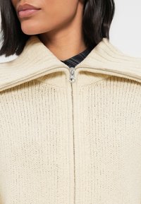 Μπεζ πουλόβερ με φερμουάρ, με ριμπ υφή, φαρδιά γιακά και μεταλλικό φερμουάρ. Κοντινή λήψη που εστιάζει στη γραμμή του λαιμού και στις λεπτομέρειες του υφάσματος.