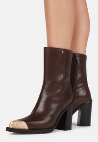 Botas de couro castanho até à metade da panturrilha, com saltos em bloco e bicos dourados metálicos, usadas sobre pernas nuas contra um fundo branco simples.