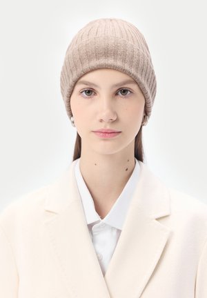 Junge Frau mit heller Haut trägt eine beige Strickmütze, silberne Creolen, ein weißes Hemd mit Kragen und einen cremefarbenen Blazer vor weißem Hintergrund.