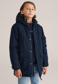 Sötétkék kapucnis dzseki, amely szövetből készült, elöl zsebekkel, cipzáros záródással és laza szabással, sötét póló fölött viselve.