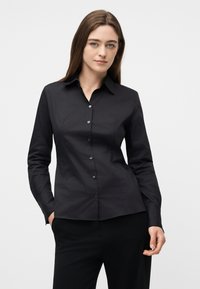 Γυναίκα με μακριά καστανά μαλλιά, που φοράει εφαρμοστό μαύρο πουκάμισο με κουμπιά και μαύρο παντελόνι, στέκεται με το ένα χέρι στην τσέπη μπροστά σε απλό φόντο.