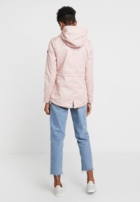 Chaqueta impermeable rosa claro con capucha, cintura ajustada y espalda más larga. Llevada sobre jeans azules con dobladillos deshilachados y zapatillas blancas.