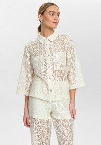 Spitzenhemdjacke in Weißbeige, mit Kragen, Knopfleiste und dreiviertellangen Ärmeln. Filigrane Auschnittmuster auf der gesamten Fläche.