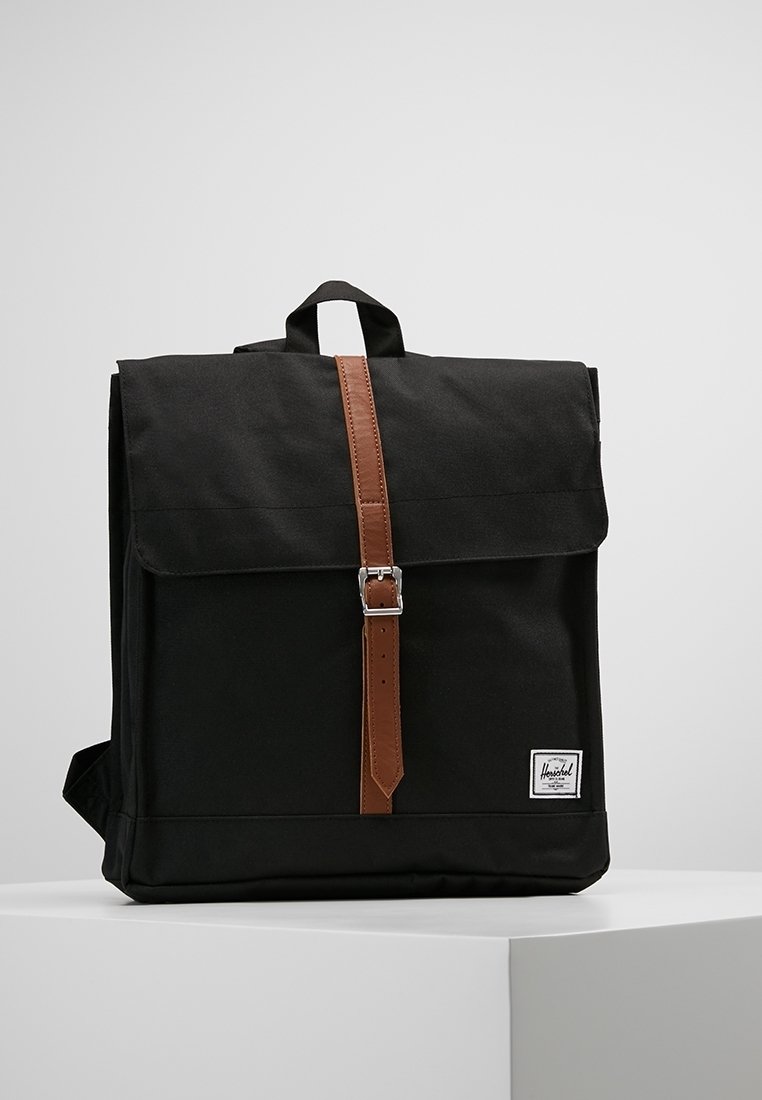 Sac à dos carré noir avec une sangle à boucle en cuir marron et un écusson logo blanc à l'avant, exposé sur un socle blanc.
