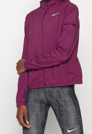 Personne portant une veste zippée Nike violette avec une capuche et un legging noir orné de logos Nike répétés en contour blanc.